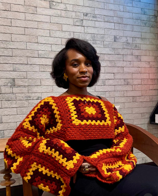 Woman wearing a colorful crochet sweater against a brick wall.