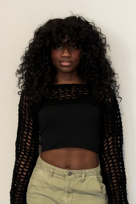 Person wearing a black crochet mesh shrug, black top, green cargo pants, and black shoes against a white wall.