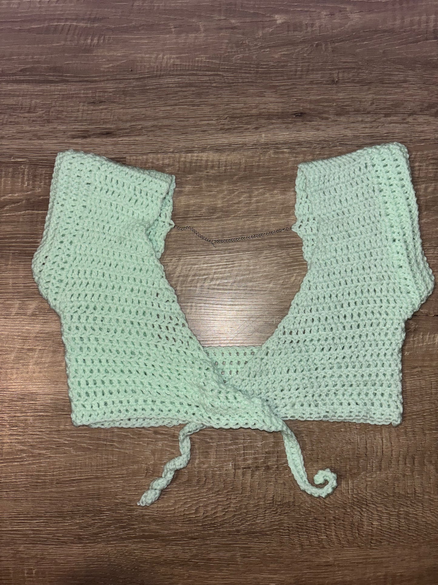 A mint green crocheted crop top with a tie-front detail, laid flat on a brown wooden surface.