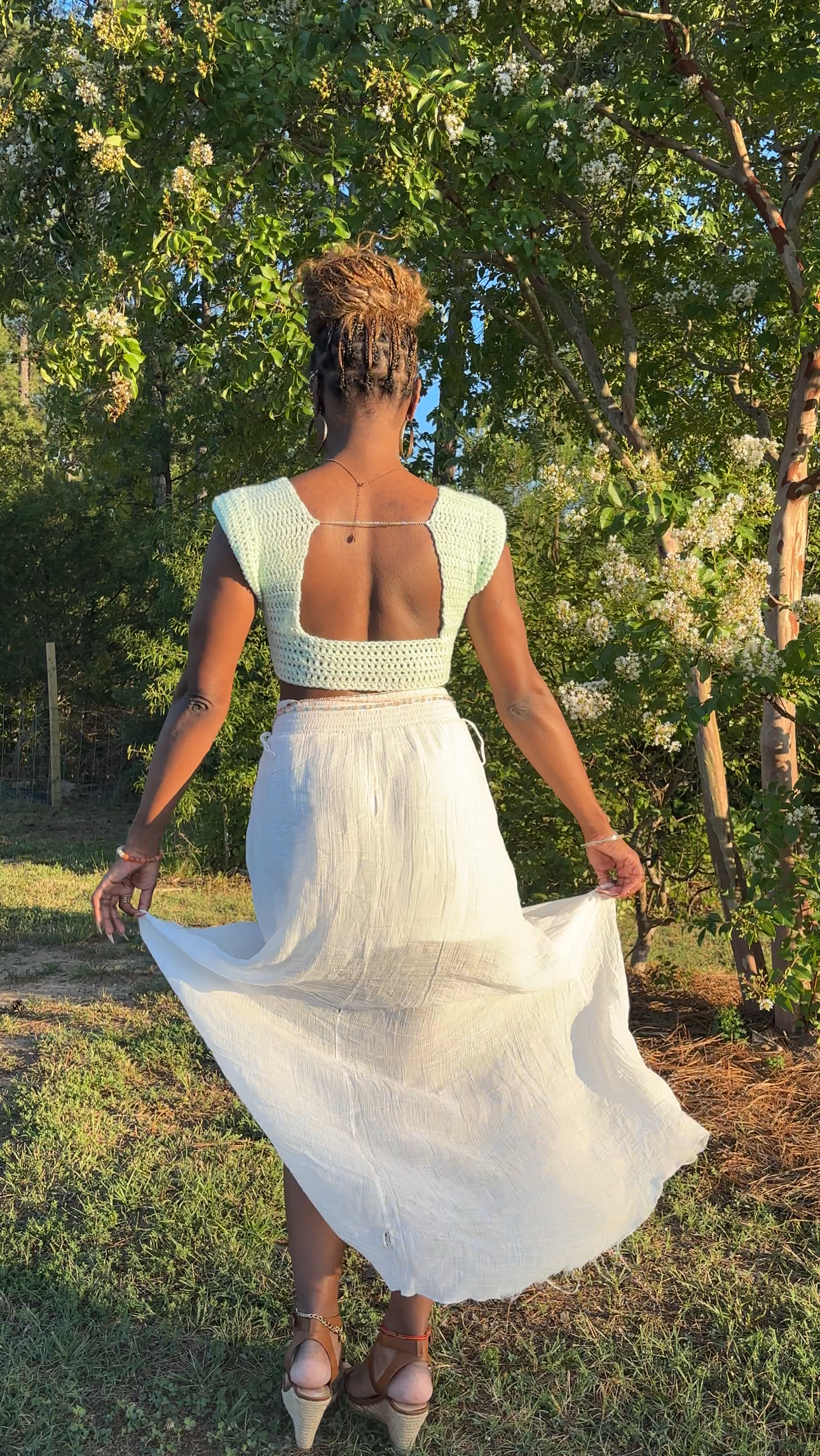 A woman stands outdoors in a garden wearing a crochet light mint green and white two-piece set with a low open back, holding out the flowing white skirt.