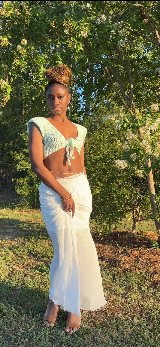 A woman stands outdoors in a grassy area with trees and flowering bushes, wearing a light mint green crochet crop top with a front tie and a flowing white maxi skirt.