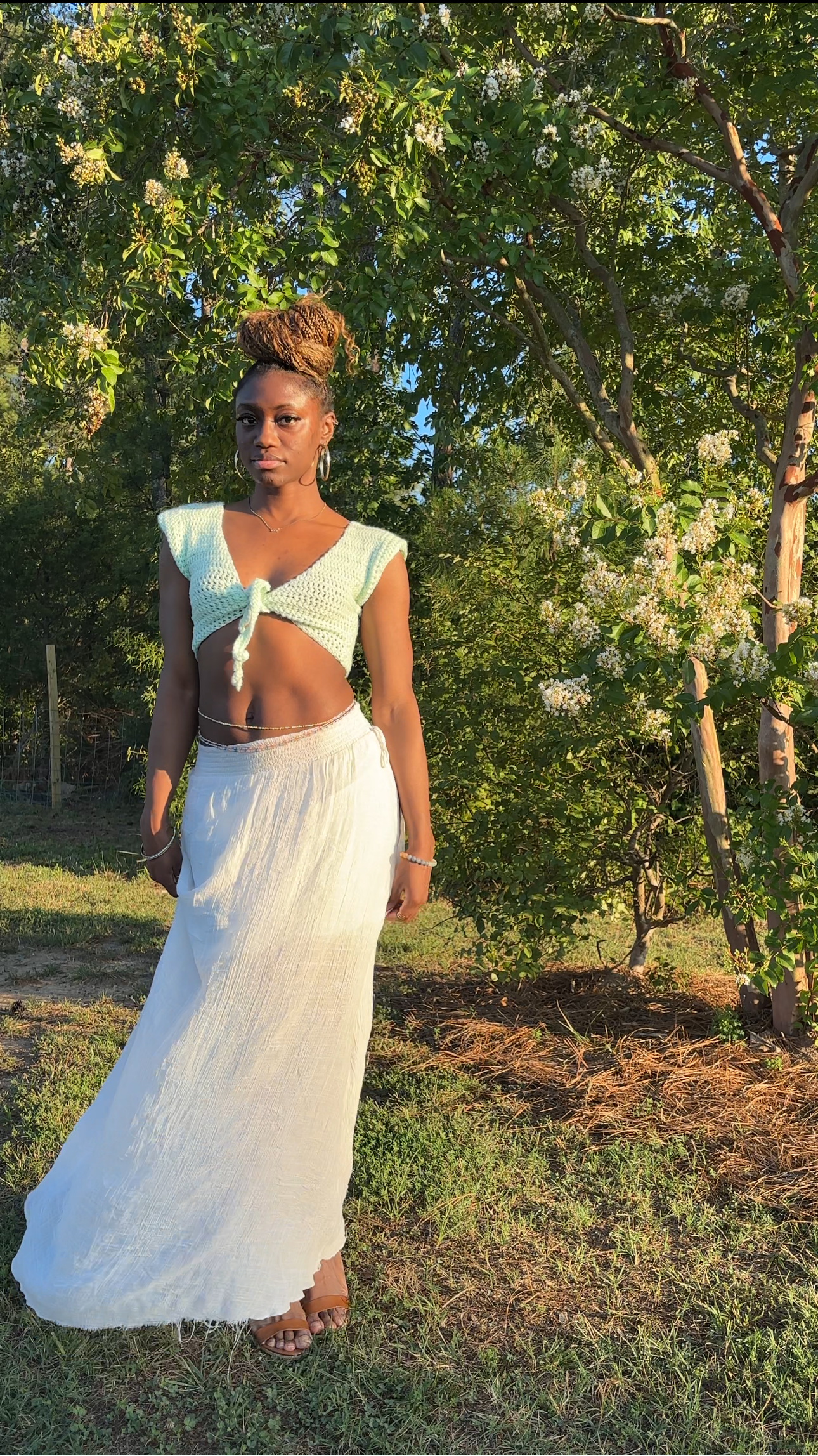 A woman wearing a white two-piece outfit consisting of a sleeveless crochet crop top with a tie detail and a white long skirt, standing outdoors in a grassy area with flowering trees in the background.