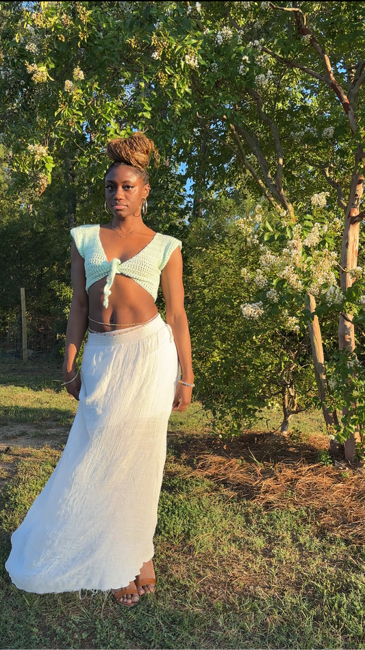 A woman wearing a white two-piece outfit consisting of a sleeveless crochet crop top with a tie detail and a white long skirt, standing outdoors in a grassy area with flowering trees in the background.