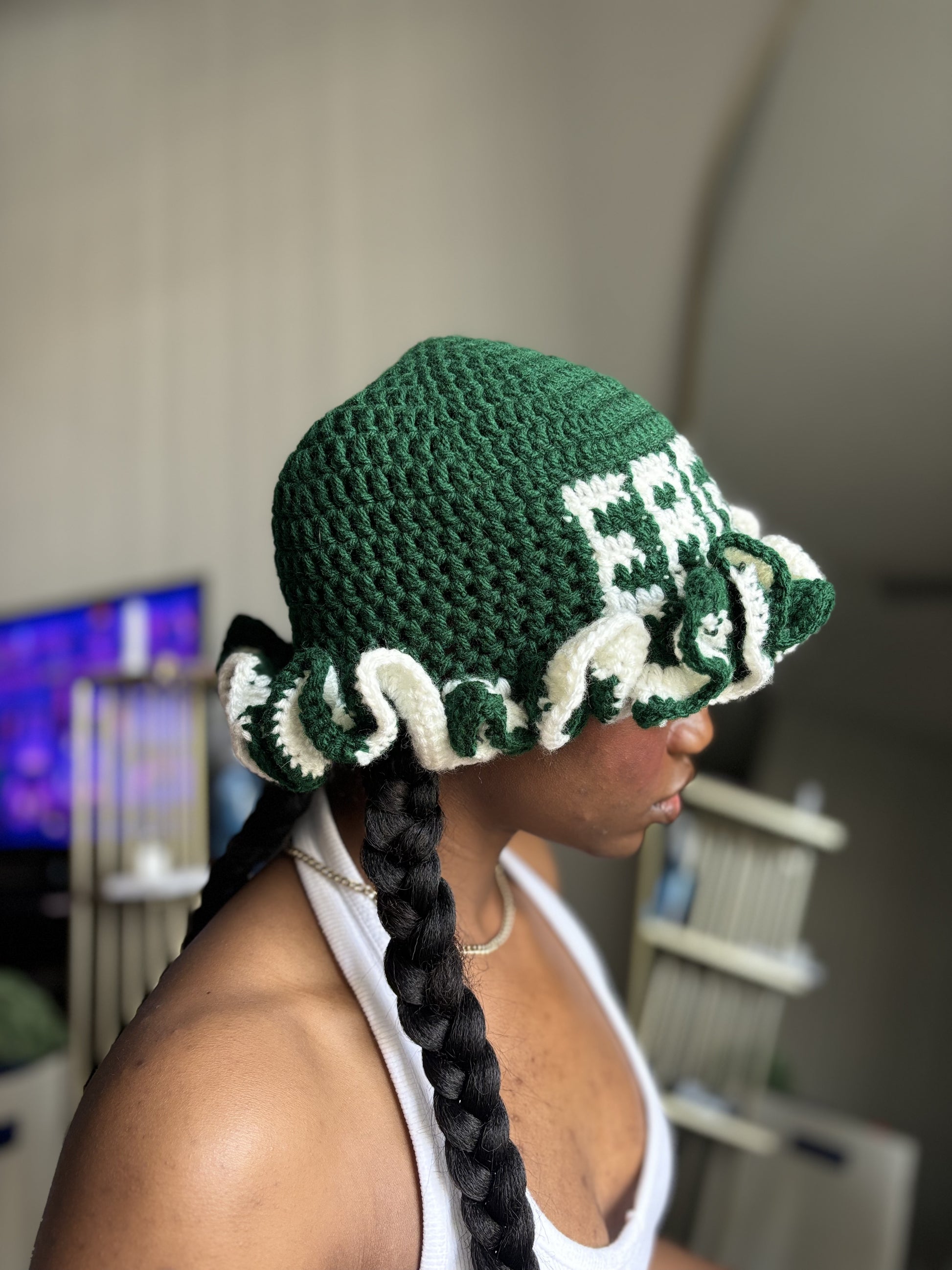 Person wearing a green and white crochet ruffle hat with braided details indoors.